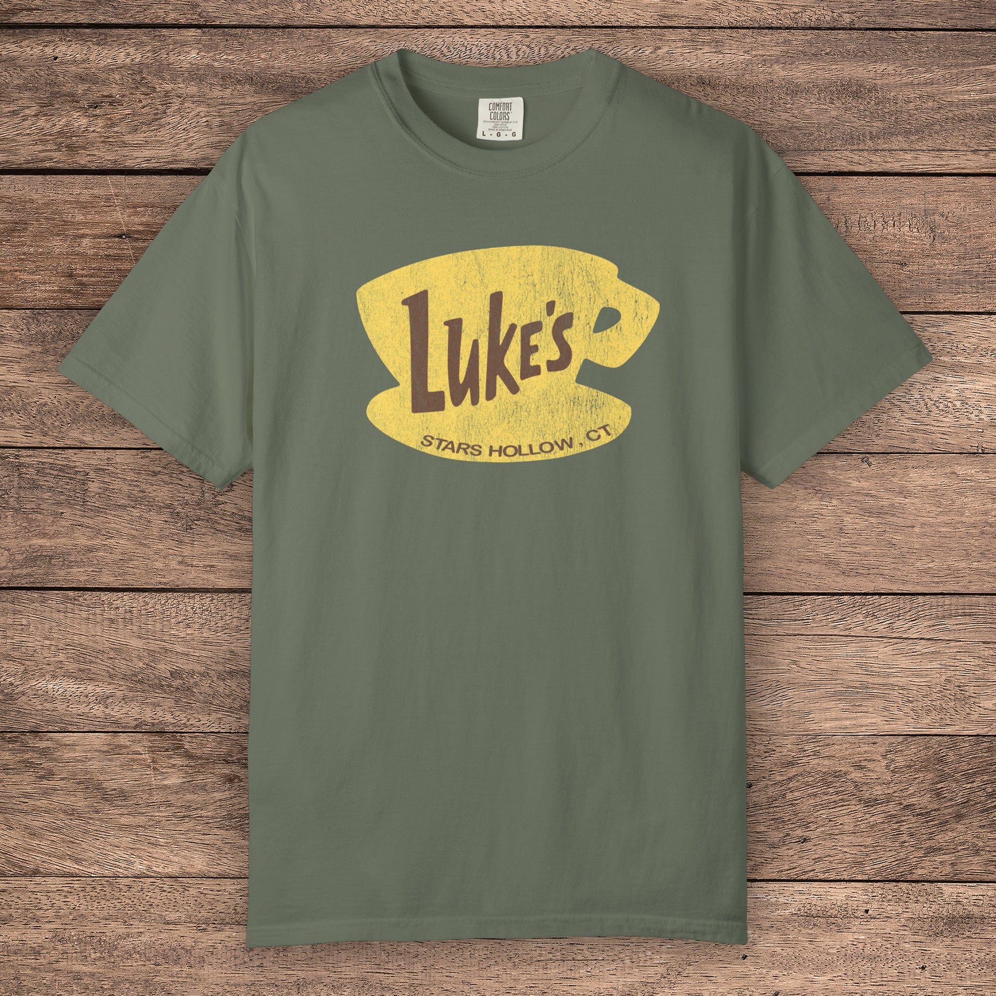 Moss green Comfort Colors t-shirt with vintage yellow Luke’s Diner logo from Gilmore Girls, laid flat on wood background.